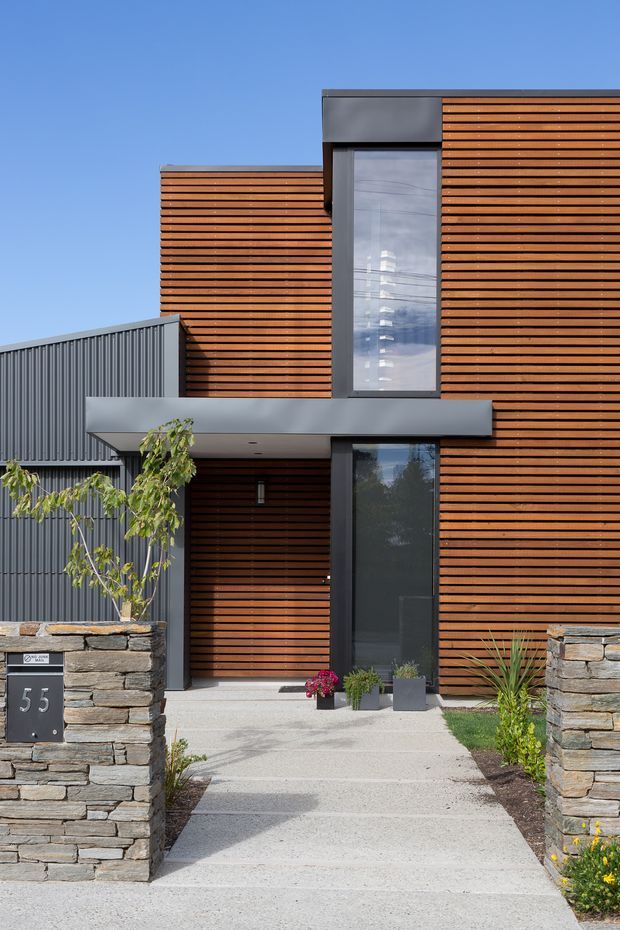 Front exterior view of a contemporary multi-level residence with strong horizontal timber slat cladding, dark grey metal wall elements, and tall vertical glazing. A covered entry canopy leads to a recessed timber-clad front door, approached by a light concrete path between stacked natural-stone boundary walls. Minimal planting and a modern letterbox complete the entry landscape.