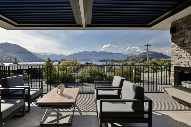 A contemporary covered outdoor terrace with a black-framed louvred roof, black metal balustrade, and a substantial stacked-stone fireplace wall. Neutral upholstered lounge chairs surround a low rectangular table, creating an outdoor seating area with expansive mountain and water views beyond.