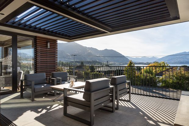 A spacious contemporary outdoor terrace or balcony furnished with a lounge seating arrangement around a low table. The terrace has dark tiled flooring, black metal balustrades, a large glazed opening, and a roofed pergola with closely spaced dark slats that cast strong linear shadows. Mountain and lake scenery are visible beyond the railing, with warm sunlight and expansive outdoor views.