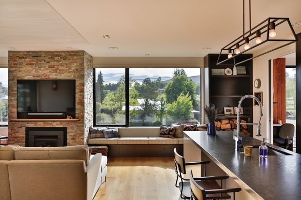 Open-plan contemporary living, dining and kitchen interior with a large island benchtop, upholstered seating, built-in window seat, dark open shelving, and a substantial brick-and-stone fireplace feature wall. Large glazing provides views to a leafy suburban landscape. A linear black metal pendant light hangs above the island.