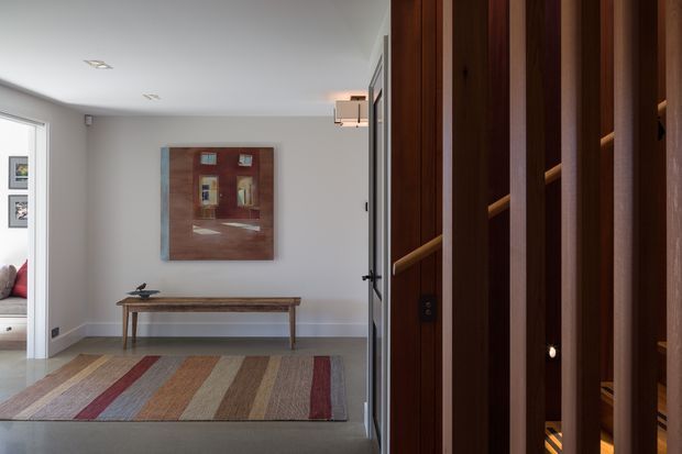 A refined residential entry hall viewed toward a wall-mounted artwork and a long timber console table. The space has pale grey walls, white trim, recessed ceiling lights, a striped area rug, and a prominent dark timber slatted architectural screen or built-in joinery at the right. A glimpse of an adjacent furnished room is visible through the opening on the left.