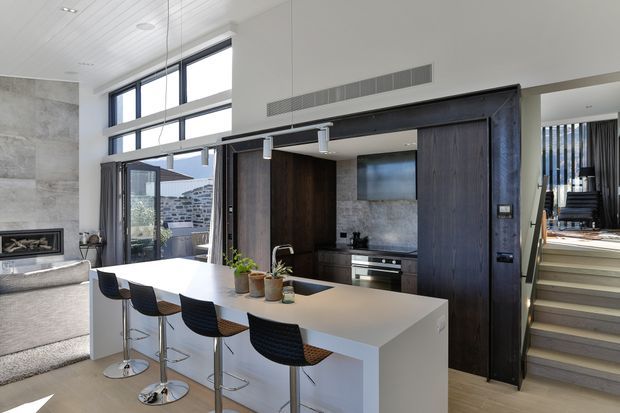 Bright open-plan contemporary kitchen with a large white island and four bar stools, dark timber cabinetry, stainless-steel appliances, and a grey stone-look splashback. The kitchen opens to a living area with a fireplace and outdoor glazing, while a staircase leads to an upper level. White panelled ceilings, high windows, and integrated linear ventilation contribute to the clean architectural character.