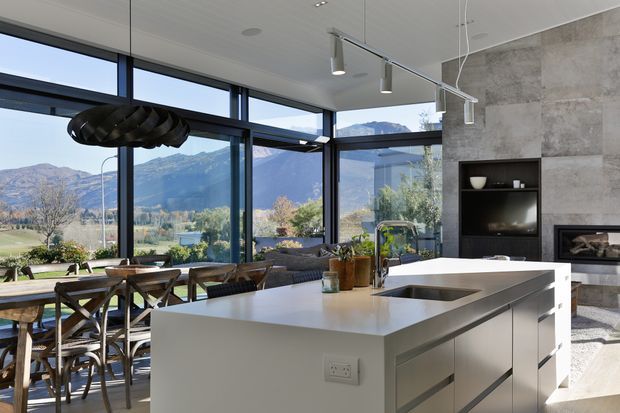 A bright contemporary open-plan kitchen and dining area with a large white island, integrated sink and induction cooktop, dark minimalist cabinetry, and a textured grey stone feature wall with fireplace. Floor-to-ceiling black-framed glazing provides expansive mountain and valley views. A dining table, sculptural pendant light, track lighting, and restrained furnishings complete the refined interior.