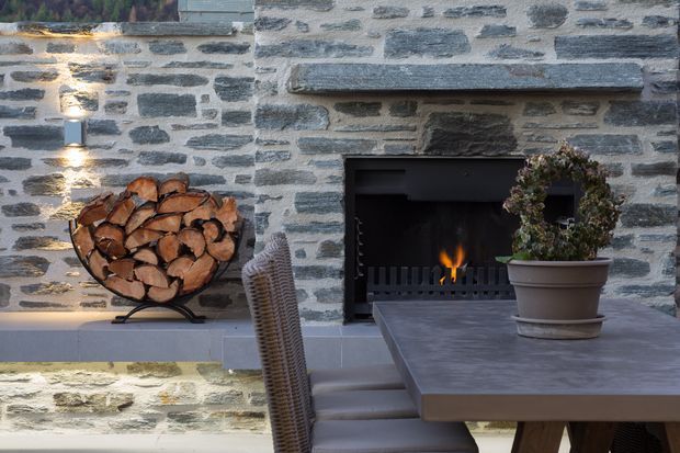 Close-up of a rustic stone-clad outdoor fireplace with a burning fire, grey stone mantel and surround, stacked firewood in a metal holder, and a large grey outdoor dining table with wicker seating. A potted wreath-like plant and wall-mounted lighting add decorative detail. The scene has a warm, rustic, refined outdoor entertaining character.