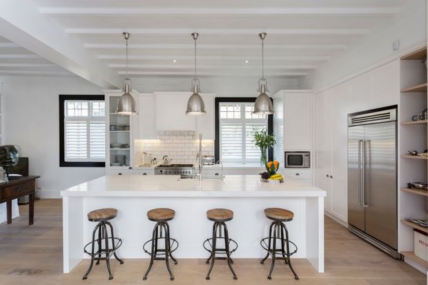 Bright, spacious white kitchen with a large central island and four vintage-style metal-and-wood stools. The room features white shaker-style cabinetry, a white subway-tile splashback, stainless-steel appliances, pale timber flooring, exposed painted ceiling beams, pendant lights, plantation shutters, and open display shelving. A small adjoining living or dining area is visible at left.