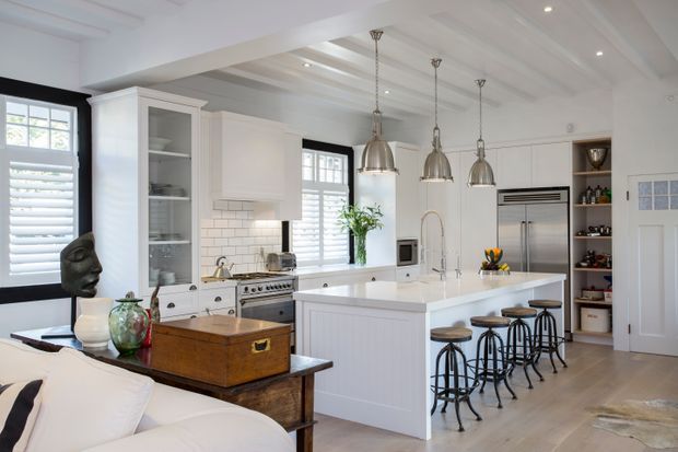 Bright, refined kitchen viewed from an adjoining living area, featuring white shaker-style cabinetry, a large central island with seating for four, stainless-steel appliances, subway-tile splashback, open shelving, pendant lights, and a coffered white ceiling. The adjacent lounge furniture and decor are partially visible in the foreground.