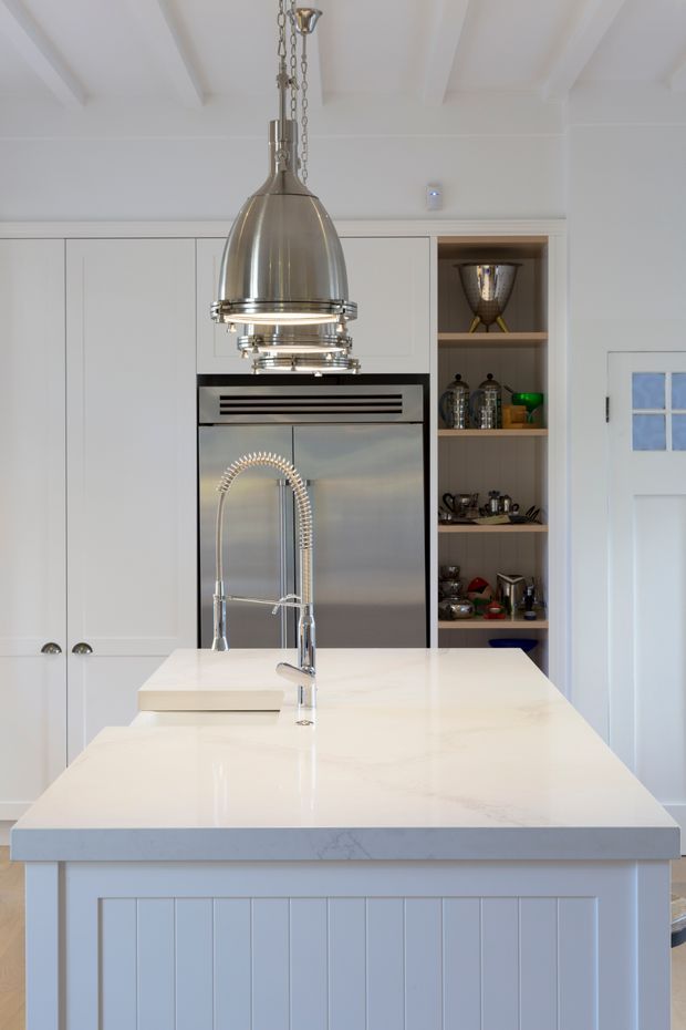 Bright, refined kitchen viewed directly toward a large island. The room features white Shaker-style cabinetry, a white stone island benchtop with integrated sink and spring faucet, stainless-steel refrigeration, open display shelving, white panelled ceiling beams, and a prominent polished metal pendant light. A white glazed door is visible at right.