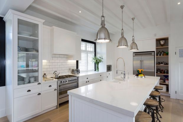 Bright, spacious white kitchen with classic shaker-style cabinetry, glass-front display storage, a subway-tile splashback, stainless-steel range and appliances, and a large central island with integrated sink and seating. Pendant lights hang from a detailed coffered ceiling, while timber flooring adds warmth.