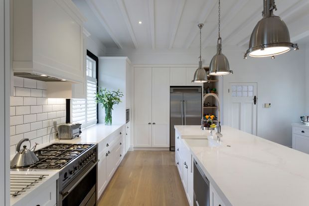 Bright, spacious white kitchen with a large central island, shaker-style cabinetry, stainless steel appliances, subway-tile splashback, timber flooring, plantation shutters, and three oversized pendant lights. The room has a refined traditional feel with abundant natural light and clean white finishes.