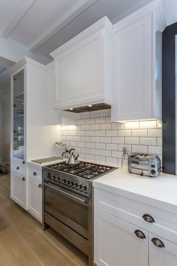 A bright, refined traditional-style kitchen with white shaker cabinetry, raised-panel doors, decorative crown moulding, integrated under-cabinet lighting, and a white subway-tile splashback. A large stainless-steel range cooker anchors the room, with a white benchtop, stainless toaster, brass-toned kettle, and light timber flooring. The compact space has a polished, classic residential fit-out.