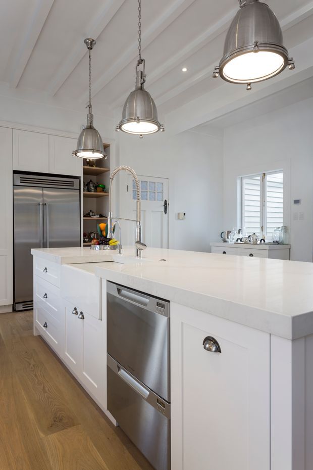 Bright, spacious white kitchen with a large island, integrated stainless-steel dishwasher, double-door refrigerator, white shaker-style cabinetry, pale engineered-stone benchtops, timber flooring, and a farmhouse-style sink with a tall spring faucet. Three oversized brushed-metal pendant lights hang from a white coffered or beamed ceiling. Open shelving, a side console, and plantation shutters add detail to the refined interior.
