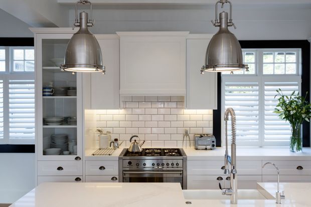 Bright, refined kitchen featuring white shaker-style cabinetry, a white subway-tile splashback, a central stainless-steel range cooker, and a large white island with double sinks and a spring-coil tap. Two oversized brushed-metal pendant lights hang above the island, while plantation shutters and black-framed windows provide contrast.