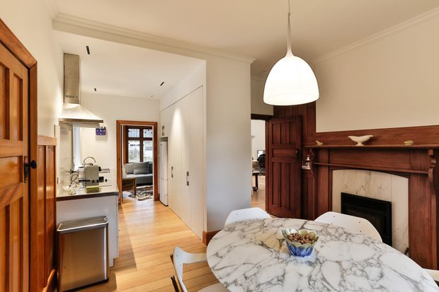 A bright open-plan dining and kitchen area in a traditional home. The foreground features a round dining table with a white marble top and contemporary white chairs, while the background contains a compact kitchen with white cabinetry, stainless-steel appliances, and timber flooring. A dark stained timber fireplace surround and mantel provide a strong heritage architectural feature, alongside timber doors and trim. A white pendant light hangs above the dining table.
