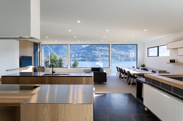 Open-plan contemporary kitchen, dining and living area with expansive windows overlooking a lake and mountainous landscape. The kitchen features light timber cabinetry, a large stainless-steel island benchtop, dark tiled flooring, integrated appliances and a black sink tap. A dining table and seating occupy the adjacent area, with a compact living zone beyond. The space is bright, clean-lined and visually connected to the scenic exterior.