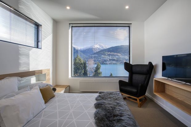 Bright contemporary bedroom with a neatly made bed, geometric grey bedding, a black upholstered lounge chair, timber bedside and media joinery, recessed ceiling lights, and large windows overlooking a lake and mountainous landscape.
