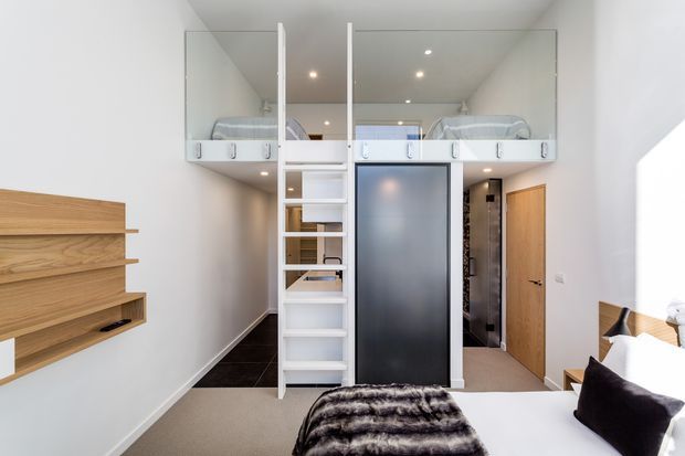Compact contemporary bedroom with a raised mezzanine sleeping platform enclosed by clear glass balustrades and accessed by a built-in white ladder. The room includes a bed in the foreground, a second bed visible on the loft level, white walls and ceiling with recessed spotlights, dark flooring on the mezzanine, a frosted-glass sliding door, timber doors, and a wall-mounted timber shelving unit.