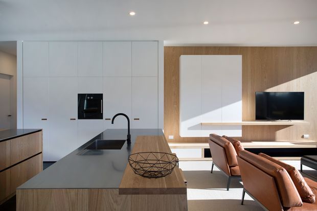 A bright, open-plan contemporary kitchen and living area. The kitchen features extensive white handleless cabinetry, a dark stone island benchtop with integrated sink and black tapware, and timber cabinetry accents. Beyond the island is a timber feature wall with a wall-mounted television, white built-in joinery, floating shelving, and several tan leather lounge chairs. Recessed ceiling lighting and strong natural light create a clean, refined atmosphere.