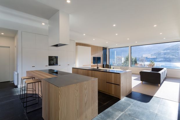 Bright open-plan contemporary kitchen and living area with a large timber island, dark stone worktops and flooring, white integrated cabinetry, and expansive corner glazing overlooking a city and mountainous landscape. Minimal recessed lighting and streamlined joinery create a clean, spacious interior.
