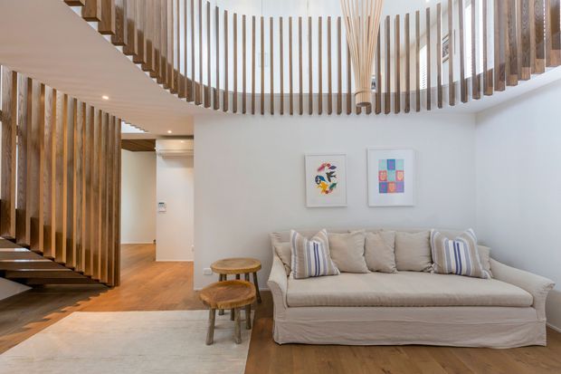 Bright contemporary living room beneath a sculptural timber staircase and curved upper-level balustrade. The room features warm timber flooring, white walls, a light upholstered sofa with striped cushions, small round timber stools, framed artwork, and integrated ceiling lighting. Vertical timber slats create a strong architectural feature around the stair and mezzanine edge.