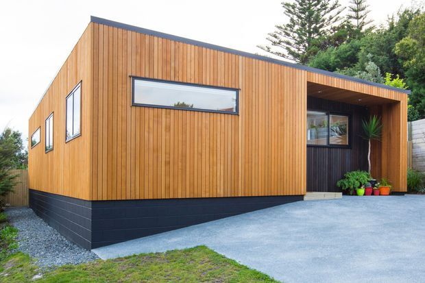 Exterior view of a compact modern single-storey building clad in vertical warm timber boards, with a dark masonry base, low-pitched roofline, black-framed windows, and a recessed covered entry porch. The structure sits beside a paved driveway and landscaped planting.