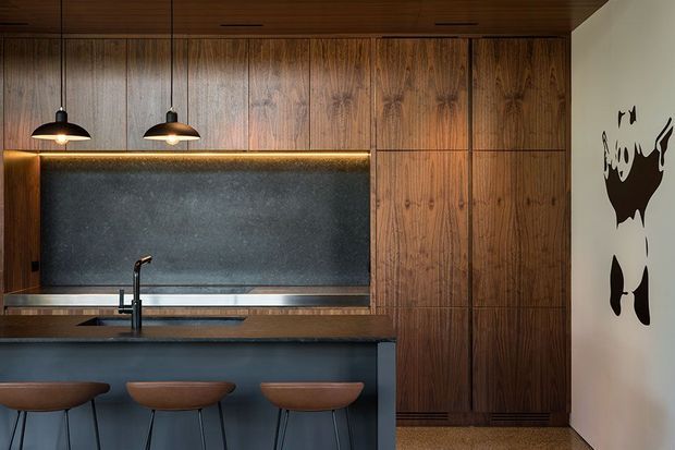 A compact, dark-toned contemporary kitchen with a long island and integrated seating. Rich timber cabinetry and timber ceiling joinery surround a charcoal stone or textured splashback, with a slim illuminated shelf detail above the benchtop. Three pendant lights hang over the island, which includes a black tap and sink. A monochrome animal wall graphic appears on the adjacent white wall.