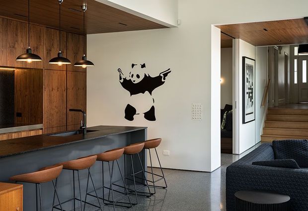 Open-plan contemporary kitchen and living area with a long dark island and timber cabinetry, lined with four leather bar stools and pendant lights. A timber-lined ceiling continues through the kitchen and adjoining entry, while a black-and-white panda mural adds a playful focal point. The living area includes a dark textured sofa and round side table, with stairs and framed artwork visible beyond.