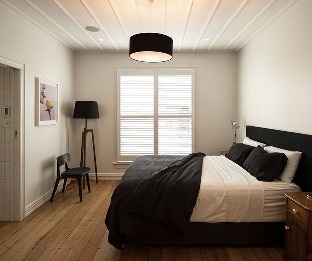 A compact, neatly styled bedroom with a black upholstered bed and dark bedding, positioned against a light neutral wall. White plantation shutters fill the central window, while a timber plank floor, black tripod lamp, framed artwork, bedside furniture, and a large black pendant light complete the room. The ceiling features white linear panel detailing and recessed lighting.