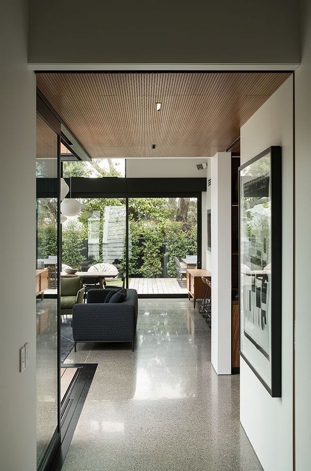A refined contemporary interior corridor opening into a living area and garden-facing outdoor deck. The space features a polished grey terrazzo floor, white walls, extensive black-framed glazing, warm timber slatted ceiling, dark built-in joinery, and understated modern furniture. The view is framed toward a leafy courtyard or garden, creating a strong indoor-outdoor connection.