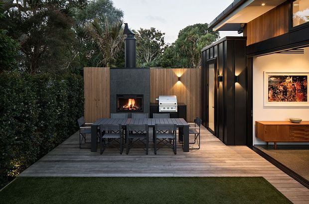 A contemporary outdoor entertaining deck beside a modern home, featuring a dark dining table with chairs, a built-in fireplace set into a textured dark wall, an outdoor barbecue, vertical timber screening, and warm architectural lighting. Large glazed openings connect the deck to an interior living area with timber cabinetry and artwork. Dense planting encloses the compact space.