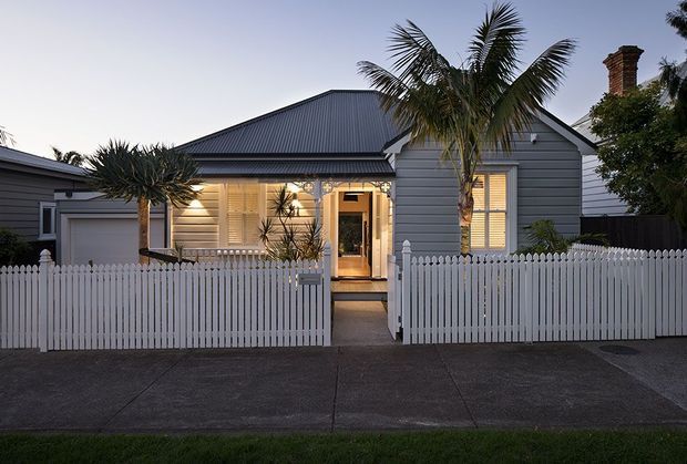 Front exterior view of a compact single-storey weatherboard house at dusk. The home has a steep corrugated metal gable roof, grey horizontal cladding, white trim, a traditional white picket fence, shuttered windows, a central entry porch, and an attached garage. Tropical palms and mature planting frame the facade.