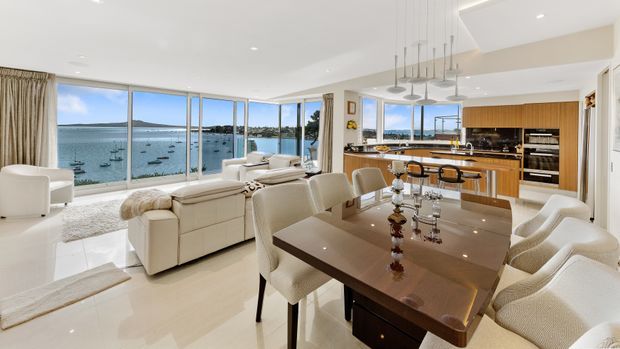 Open-plan waterfront interior combining a dining area, living room and modern kitchen. The space features extensive floor-to-ceiling glazing with a harbour view, pale polished flooring, cream upholstered furniture, a large dark timber dining table, and warm timber kitchen cabinetry. A sculptural linear pendant hangs above the dining area.