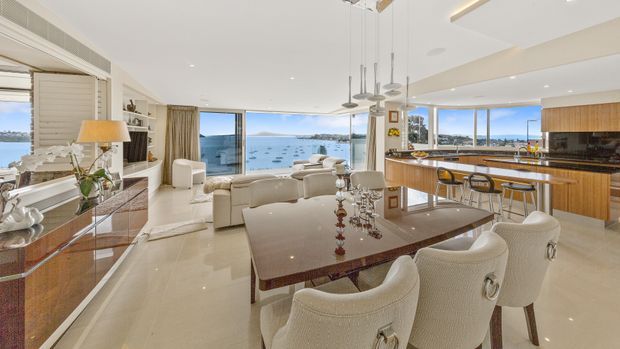Bright, open-plan coastal apartment interior combining a dining area, living room and kitchen, with expansive glazing and views over the water. The space features glossy light flooring, cream upholstered seating, a dark stone dining table, warm timber kitchen cabinetry, a large island with bar seating, and contemporary pendant lighting.
