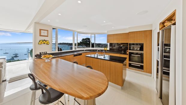 Bright, spacious contemporary kitchen with a large curved timber island and integrated seating, warm wood cabinetry, dark stone benchtops, stainless-steel appliances, and extensive curved glazing overlooking a waterfront setting.