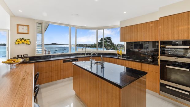 A spacious contemporary kitchen with warm timber cabinetry, glossy black stone benchtops and splashback, a central island with sink, integrated stainless-steel appliances, and expansive curved glazing overlooking a coastal waterfront landscape.