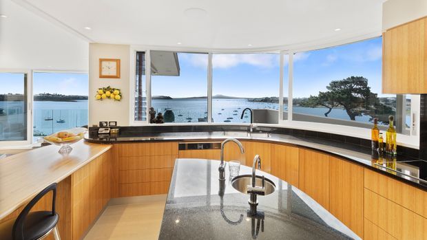Bright contemporary kitchen with extensive curved bay windows overlooking a calm coastal bay. The space features warm timber cabinetry, dark polished stone benchtops and splashback surfaces, a central island with integrated sinks and taps, and a dining nook at left. Abundant glazing creates an airy, open connection to the waterfront setting.