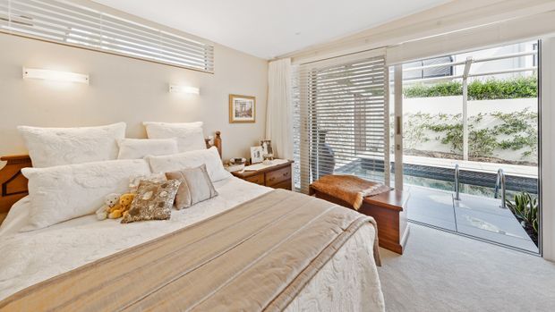 Bright, refined bedroom with a large upholstered bed dressed in layered white and beige bedding, bedside tables, wall lights, and a timber dresser. Full-height glazed doors with horizontal blinds open toward a small outdoor courtyard or lightwell, bringing in abundant natural light. The room has a neutral palette, soft textures, and a calm, comfortable feel.