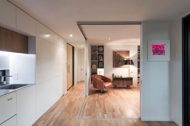 A contemporary open-plan apartment interior viewed from the kitchen toward a living room. The kitchen has extensive white flat-panel cabinetry, a stainless-steel sink and integrated appliances, with a timber-look vertical feature panel. Warm timber flooring continues through the space, separated from the living area by a large black-framed glazed sliding partition. The living room contains a brown leather armchair, a low timber media console, open shelving, artwork and a floor lamp. The palette is warm and neutral with subtle black accents.