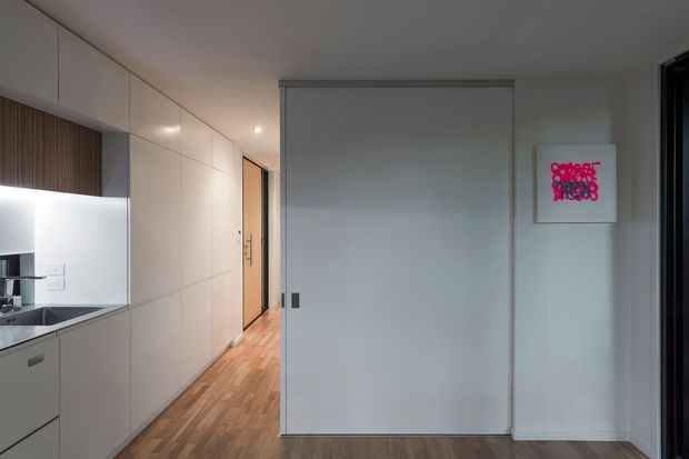 Compact contemporary kitchen viewed toward a hallway, with full-height pale cabinetry, integrated appliances, a dark benchtop, timber flooring, and a large plain sliding door or partition. Minimal white walls and recessed lighting create a clean, understated interior, with a small framed artwork adding a bright pink accent.