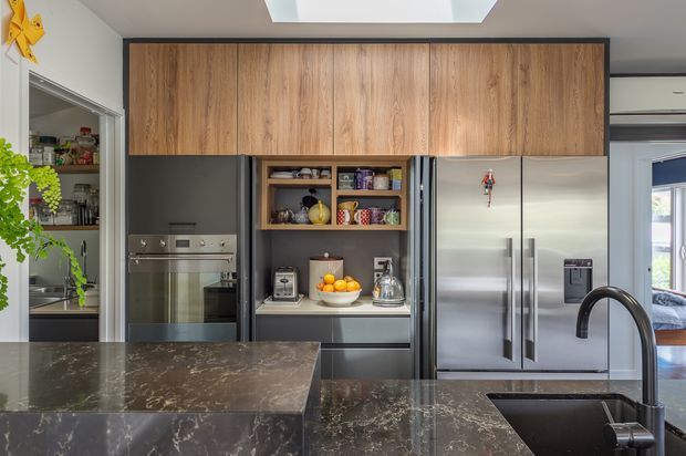 A contemporary kitchen with dark stone benchtops, integrated stainless-steel appliances, a large stainless refrigerator, and extensive warm timber overhead cabinetry. Open shelving displays kitchenware and pantry items, while a skylight brings natural light into the space. A walk-in pantry is visible to the left and an adjoining room is visible through the doorway on the right.