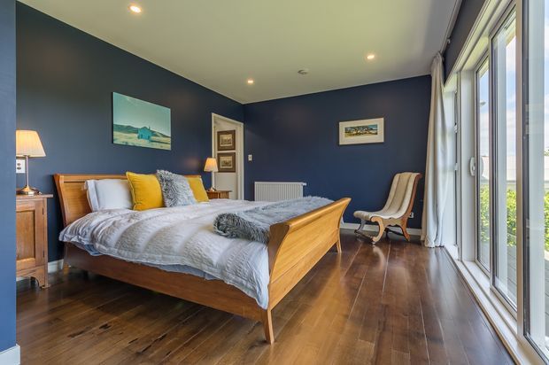 A well-appointed bedroom with deep navy-blue walls, warm timber flooring, and a mid-century-style timber bed with a curved footboard. The bed is dressed in white and grey bedding with mustard and pale blue accent cushions. A sculptural lounge chair sits beside full-height glazed doors and windows, bringing in abundant daylight and views of greenery. Recessed ceiling lights, framed artwork, bedside tables, and a radiator complete the room.