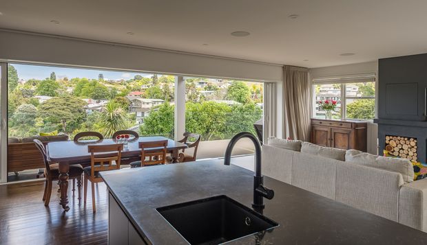 Open-plan contemporary living and dining area viewed from a kitchen island. The foreground features a dark stone island benchtop with an undermount sink and curved black tap. Beyond it are a timber dining table and chairs, a pale upholstered sofa, timber cabinetry, and a dark fireplace surround with stacked firewood. Large floor-to-ceiling windows provide leafy suburban views and abundant natural light. The room has polished dark timber flooring, neutral walls, recessed ceiling lights, and full-height curtains.