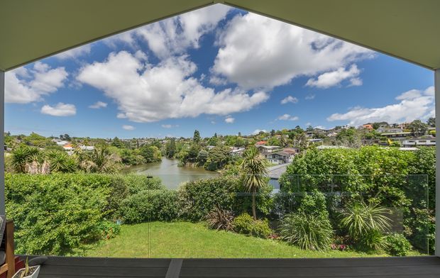 A wide view from an elevated covered outdoor space overlooking a lush landscaped garden, a small lake or pond, and a suburban neighbourhood. The opening is framed by a dark roof or ceiling structure, with clear glass balustrading across the foreground and dark timber-like decking below. Bright blue sky and scattered clouds create an airy outlook.
