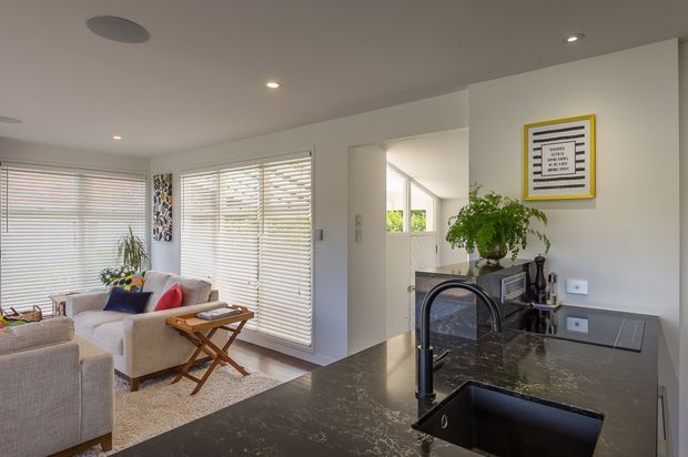Open-plan interior view from a kitchen island toward a compact living room. The kitchen has a dark stone benchtop with an undermount sink and curved black tap, while the adjacent living area contains light upholstered seating, a timber folding tray table, patterned cushions and a pale rug. White walls, recessed ceiling lights, wide horizontal blinds and a framed graphic artwork create a clean, bright contemporary setting. A planted sideboard is visible beyond the kitchen opening.