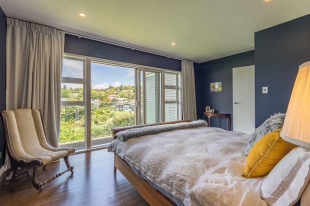 A contemporary bedroom with a large bed dressed in white textured bedding and grey throws, dark navy-blue walls, full-height curtains, and expansive sliding windows overlooking a leafy residential landscape. A sculptural upholstered rocking chair sits beside the window, while a yellow accent cushion and warm table lamp add colour and softness. Recessed ceiling lights and dark timber flooring complete the room.