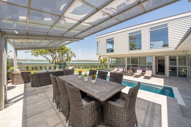Bright contemporary outdoor entertaining area beside a narrow swimming pool, featuring a large wicker dining setting, lounge seating, and a transparent covered pergola. The space adjoins a two-storey house with light horizontal cladding, extensive glazing, and a paved terrace.