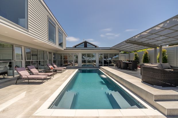 A bright contemporary courtyard centered on a long rectangular swimming pool, surrounded by pale paved terraces, lounge chairs, and outdoor seating. The multi-wing home has light horizontal cladding, extensive glazing, and a large translucent covered outdoor entertaining area with integrated planting.