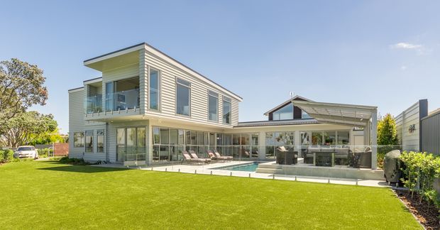 A bright exterior view of a contemporary multi-level home with light horizontal weatherboard cladding, a low-pitched roof, extensive glazing, and a covered outdoor entertaining area. The house opens onto a broad manicured lawn with a small swimming pool, outdoor seating, and a sheltered kitchen or barbecue zone.