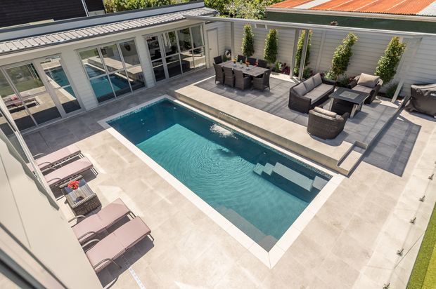 Elevated view of a landscaped residential courtyard centered on a rectangular swimming pool, surrounded by pale tiled paving, sun loungers, outdoor dining furniture, and a separate outdoor seating area. The courtyard is enclosed by light grey horizontal weatherboard walls with extensive glazed doors and windows, climbing greenery, and a corrugated metal roof visible beyond.
