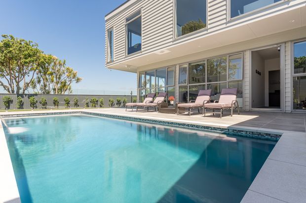 Bright exterior view of a contemporary two-storey home beside a swimming pool. The building has pale horizontal weatherboard-style cladding, large glazed openings, a projecting upper floor, and a light paved pool terrace furnished with lounge chairs.