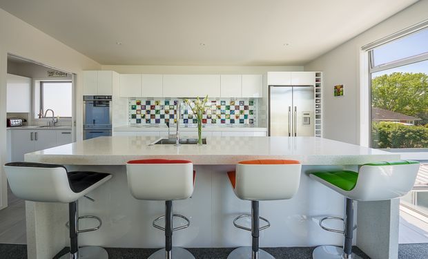 Bright contemporary kitchen with a large white island and integrated breakfast bar, four modern sculptural bar stools, extensive white cabinetry, stainless-steel appliances, and a colourful mosaic tile splashback. Large windows provide abundant natural light and views of greenery outside.